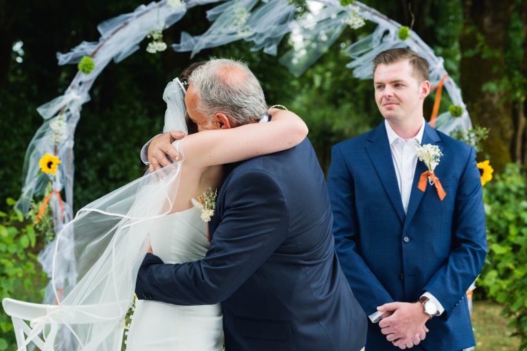 Une mariée vêtue d'une robe blanche et d'un voile serre dans ses bras un homme plus âgé en costume bleu marine près d'une arche décorée en plein air. Un homme en costume bleu, avec une boutonnière et un ruban orange se tient à droite, observant et souriant légèrement. Des arbres verts et des fleurs se trouvent à l'arrière-plan.