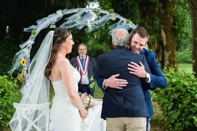 Une mariée en robe blanche et voile sourit tandis que le marié, en costume bleu, étreint un homme plus âgé. Ils se tiennent à l'extérieur, sous une arche de tissu blanc et de fleurs. Un homme portant une médaille se tient derrière une table, officiant la cérémonie. De la verdure entoure la scène.
