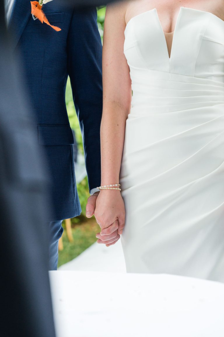 Une mariée vêtue d'une robe blanche sans bretelles avec des détails froncés et un marié vêtu d'un costume bleu foncé se tiennent par la main. La mariée porte un bracelet de perles. Leurs visages ne sont pas cadrés et ils se tiennent à l'extérieur, peut-être lors d'une cérémonie de mariage.