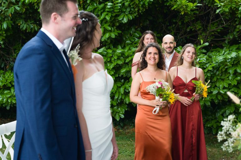 Une mariée en robe bustier blanche et un marié en costume bleu marine sourient en plein air ; derrière eux, quatre demoiselles d'honneur en robe orange brûlé tiennent des bouquets et sourient, tandis que des buissons verts luxuriants forment l'arrière-plan.