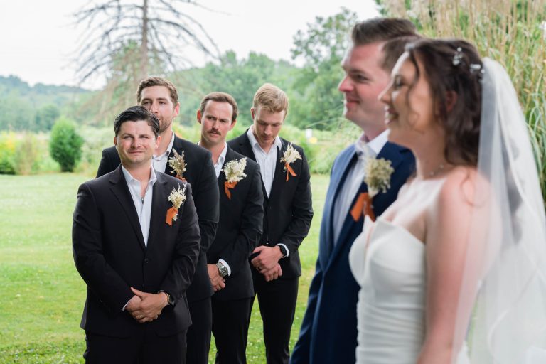 Une mariée et un marié se tiennent face à face en plein air lors d'une cérémonie de mariage. Derrière eux, quatre garçons d'honneur en costume noir avec des boutonnières et des rubans orange les regardent en souriant. De l'herbe verte, des arbres et des plantes sont visibles à l'arrière-plan.