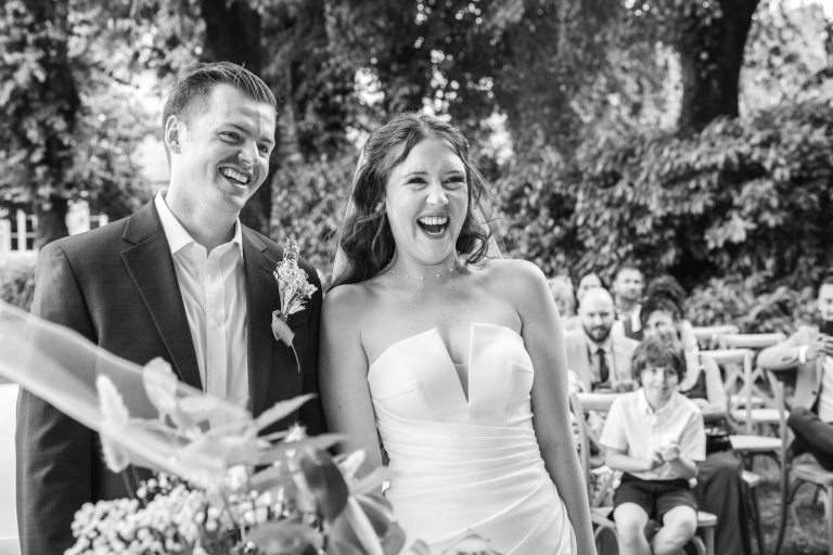 Une mariée souriante en robe bustier et un marié en costume se tiennent côte à côte en plein air, riant joyeusement, avec des arbres feuillus et des invités assis en arrière-plan. L'atmosphère est lumineuse et festive.