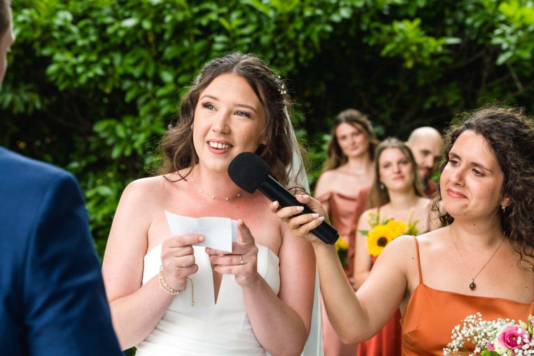 Une mariée en robe blanche tient un papier et sourit tout en parlant dans un microphone tenu par une demoiselle d'honneur en robe orange. Plusieurs demoiselles d'honneur portant des robes assorties et tenant des fleurs se tiennent à l'arrière-plan. De la verdure luxuriante entoure le groupe.