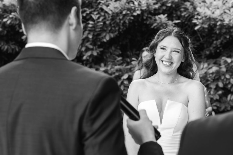 Une mariée vêtue d'une robe blanche sans bretelles sourit joyeusement à une personne en costume tenant un microphone, probablement lors d'une cérémonie de mariage, avec de la verdure en arrière-plan. L'image est en noir et blanc.
