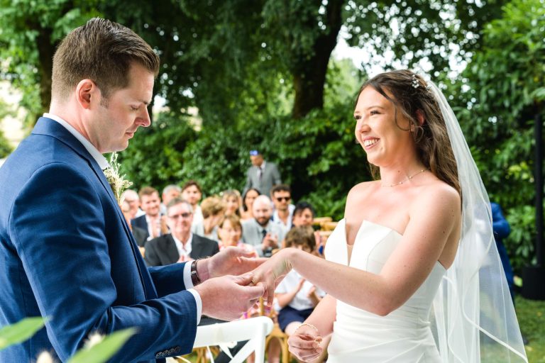 Un marié en costume bleu passe la bague au doigt de la mariée, qui sourit de toutes ses dents. Ils se tiennent en plein air, entourés de verdure luxuriante et d'invités assis qui assistent à la cérémonie de mariage. La mariée porte une robe blanche sans bretelles et un voile.