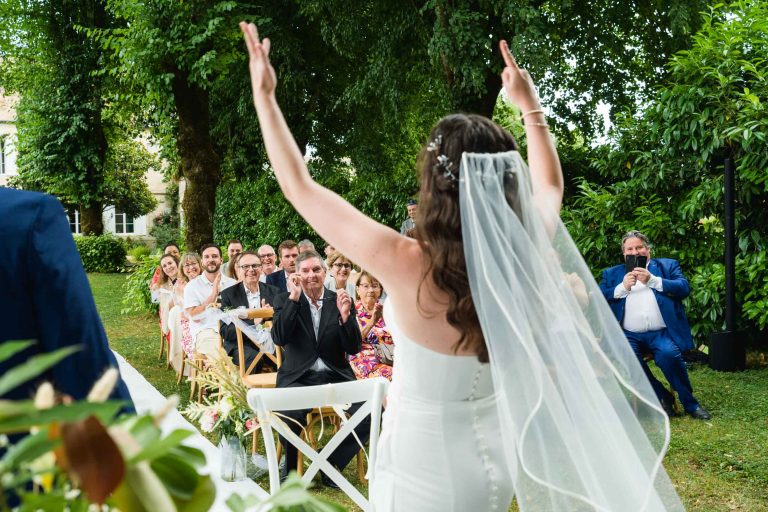 Une mariée vêtue d'une robe blanche et d'un voile, vue de dos, les bras levés, fait face à des invités assis qui l'acclament lors d'une cérémonie de mariage en plein air. Les invités souriants sont assis sur des chaises en bois sous des arbres verdoyants, et un homme en costume bleu prend une photo.