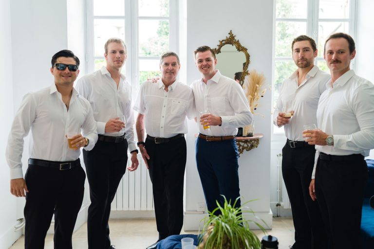 Six hommes vêtus de chemises blanches et de pantalons sombres se tiennent à l'intérieur, posant et souriant, chacun tenant un verre. L'un d'eux porte des lunettes de soleil, un autre une moustache. Ils se trouvent dans une pièce lumineuse avec de grandes fenêtres, un miroir encadré d'or et une table avec des fleurs séchées en arrière-plan.