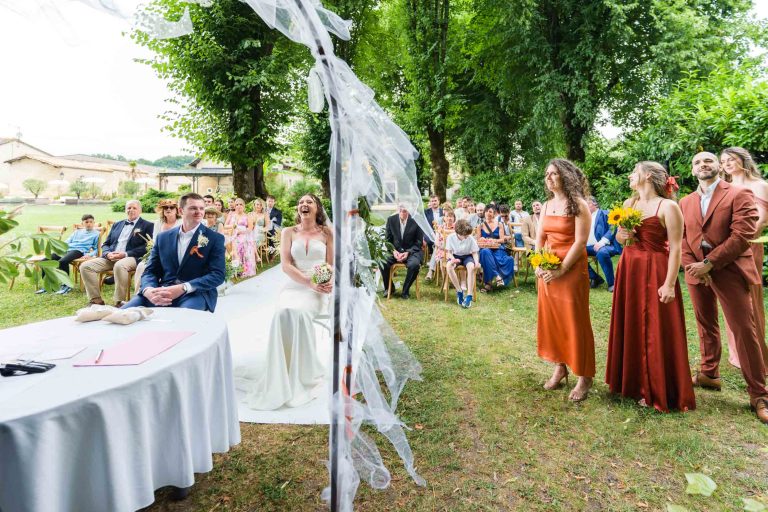 Une cérémonie de mariage en plein air montre une mariée en robe blanche riant à côté d'un marié en costume bleu. Des invités sont assis sur des chaises à l'arrière-plan. Trois demoiselles d'honneur en robe orange rouille tiennent des tournesols, à côté d'un garçon d'honneur en costume marron. Des arbres encadrent la scène.