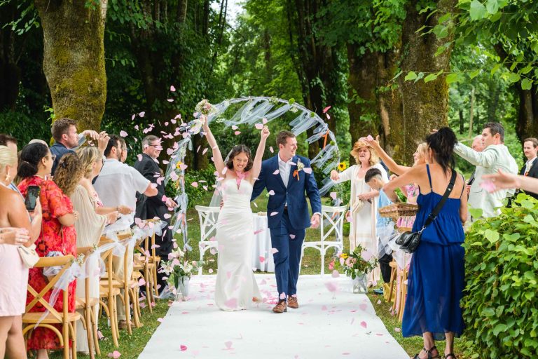 Des mariés joyeux descendent une allée en plein air, entourés d'invités enthousiastes qui lancent des pétales de fleurs. La mariée lève les deux bras en souriant, tandis que le marié lui tient la main. Des arbres verdoyants et une arche décorative encadrent cette scène de mariage animée et festive.