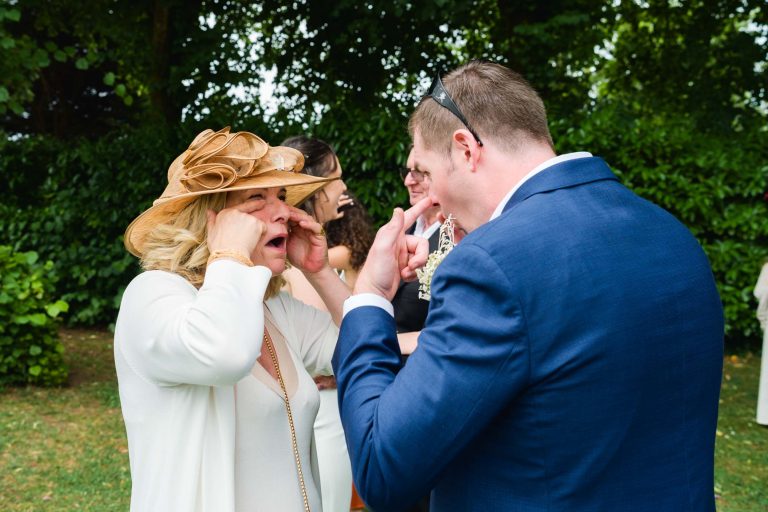 Une femme vêtue d'une tenue beige et d'un chapeau décoratif et un homme vêtu d'un costume bleu se tirent la bourre en étirant la peau de leurs yeux avec leurs doigts. Ils sont en plein air, sur de l'herbe, avec des arbres et plusieurs autres personnes floues en arrière-plan, et jouent ensemble.