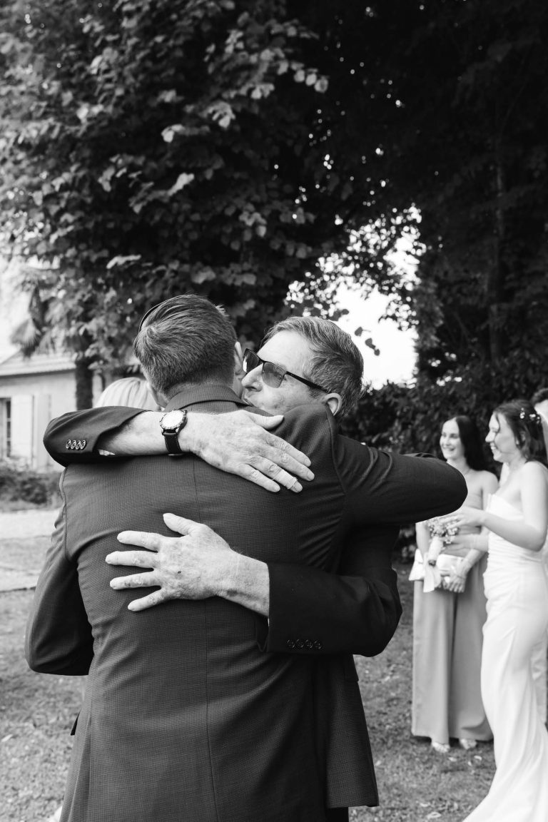 Deux hommes en costume s'embrassent chaleureusement en plein air, l'un faisant face à la caméra et portant des lunettes de soleil. À l'arrière-plan, trois femmes en robe de soirée se tiennent debout et sourient, des arbres et un bâtiment étant partiellement visibles. L'image est en noir et blanc.
