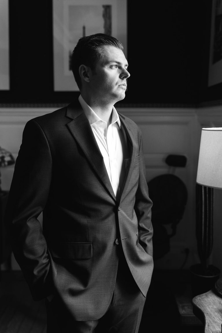 Un homme en costume se tient à l'intérieur, regardant hors du cadre vers la droite avec une expression sérieuse. Il a une main dans sa poche et est éclairé par une lumière douce et naturelle. L'arrière-plan comprend des œuvres d'art encadrées, des panneaux de bois et une lampe. La photo est en noir et blanc.