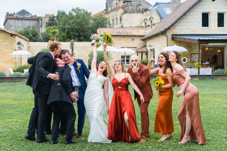 Une joyeuse fête de mariage pose sur l'herbe à l'extérieur d'un bâtiment rustique. La mariée en blanc et les demoiselles d'honneur en robes de satin s'amusent avec des bouquets, tandis que les garçons d'honneur en costume rient et se serrent dans les bras. Le groupe rayonne d'excitation et de célébration par une journée lumineuse et nuageuse.