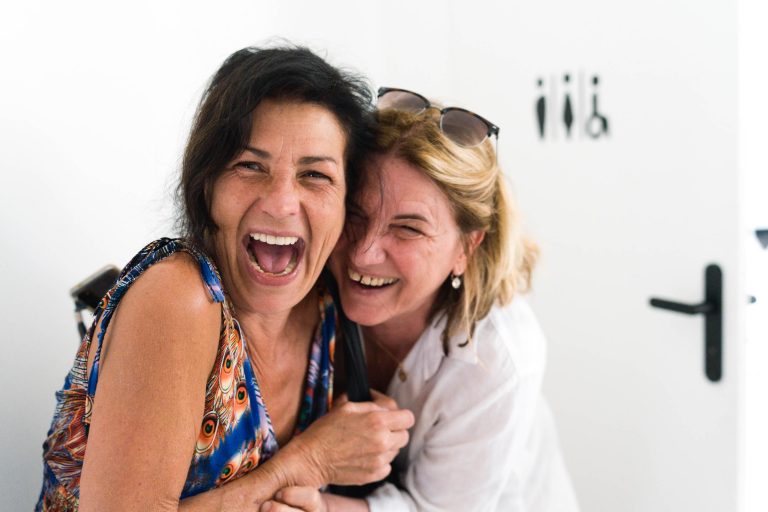 Deux femmes rient joyeusement dans une pièce lumineuse. L'une a les cheveux noirs et porte un haut sans manches coloré ; l'autre a les cheveux plus clairs, des lunettes de soleil sur la tête et une chemise blanche. Elles s'enlacent étroitement, souriant largement près d'un mur blanc et d'une porte noire.