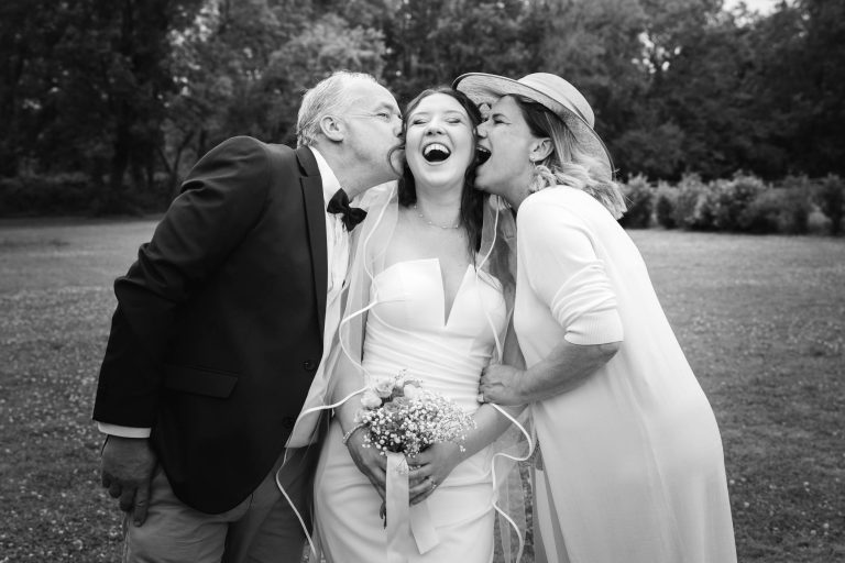 Une mariée vêtue d'une robe blanche et d'un voile se tient en plein air, tenant un bouquet et riant joyeusement tandis qu'un homme et une femme plus âgés, probablement ses parents, l'embrassent chacun sur une joue. Le trio se tient près l'un de l'autre, entouré de verdure. La photo est en noir et blanc.