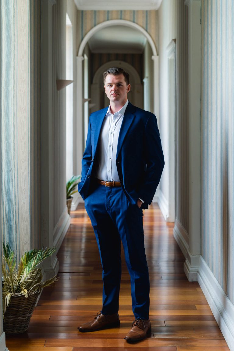 Un homme vêtu d'un costume bleu, d'une chemise blanche et de chaussures marron se tient dans un couloir lumineux au sol en bois et au papier peint rayé. Il a les mains dans les poches et une expression sérieuse. Des plantes en pot sont disposées le long des murs du couloir.