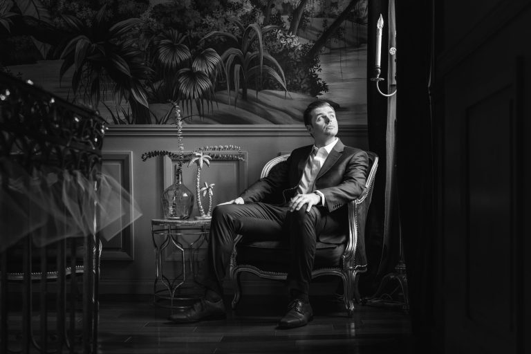 Un homme en costume est assis sur un fauteuil orné, regardant pensivement vers une applique murale allumée. Il se trouve dans une pièce somptueuse avec un sol en bois sombre, une table d'appoint tenant un vase en verre et une scène de forêt tapissée en arrière-plan. L'image est en noir et blanc.