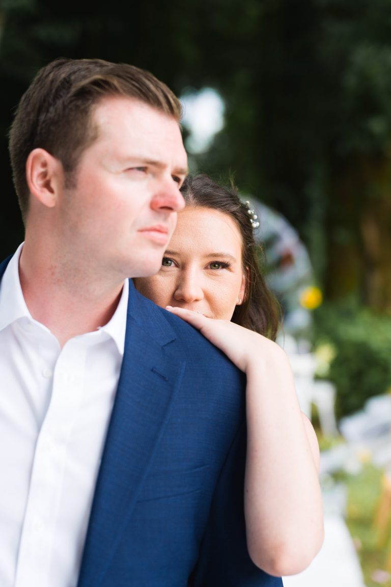 Une femme se tient derrière un homme en costume bleu, posant sa main sur son épaule et regardant la caméra. L'homme regarde sur le côté. Tous deux sont à l'extérieur, avec de la verdure et des décorations blanches floues en arrière-plan.
