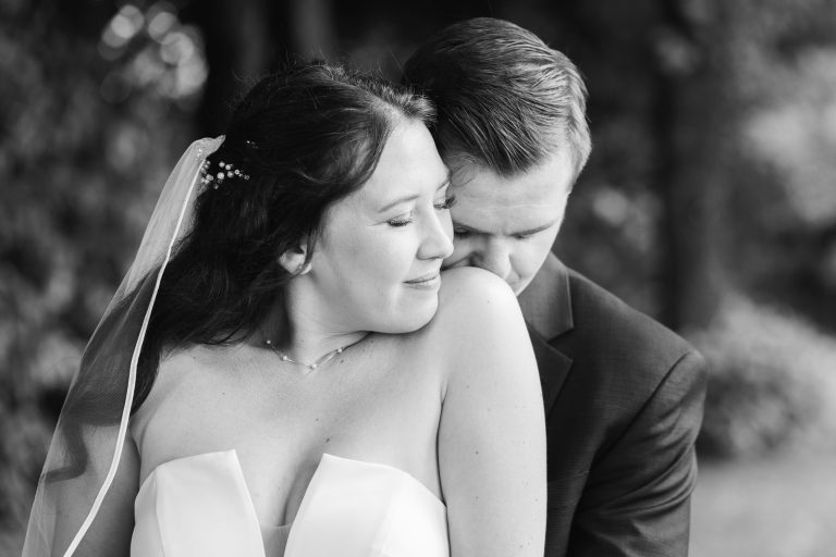 Photo en noir et blanc d'une mariée et d'un marié. Le marié, en costume, enlace la mariée par derrière et lui embrasse doucement l'épaule. La mariée sourit doucement, les yeux fermés, avec un voile et des épingles à cheveux en perles. De la verdure floue se trouve à l'arrière-plan.