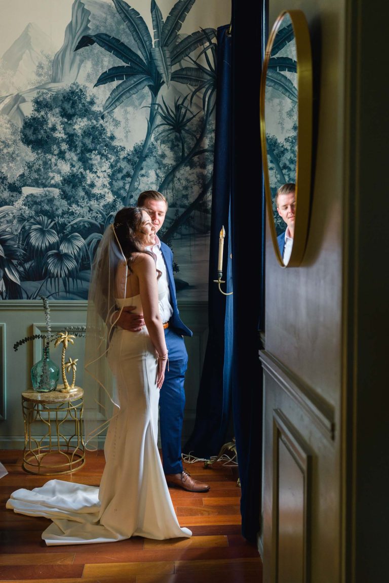 Les mariés se tiennent l'un à côté de l'autre dans une salle élégante aux rideaux bleu foncé et ornée d'une peinture murale tropicale. La mariée porte une robe et un voile blancs ; le marié porte un costume bleu. Leur reflet est visible dans un miroir rond fixé au mur.