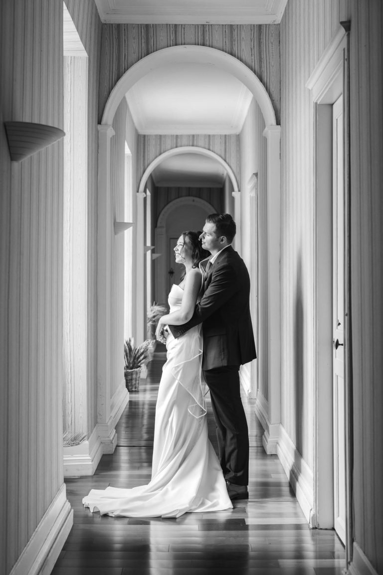 Les mariés se tiennent dans un couloir ensoleillé aux portes cintrées. Le marié étreint la mariée par derrière alors qu'ils regardent tous deux par la fenêtre. La mariée porte une longue robe fluide et le marié est en costume. La scène est en noir et blanc.