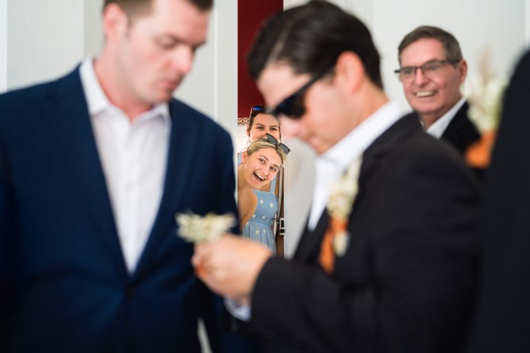 Une femme en robe bleue sourit et se penche dans l'embrasure d'une porte derrière trois hommes en costume, dont l'un, flou, ajuste une boutonnière, tandis qu'un autre homme plus âgé, portant des lunettes, sourit à l'arrière-plan. L'atmosphère est animée et joyeuse.