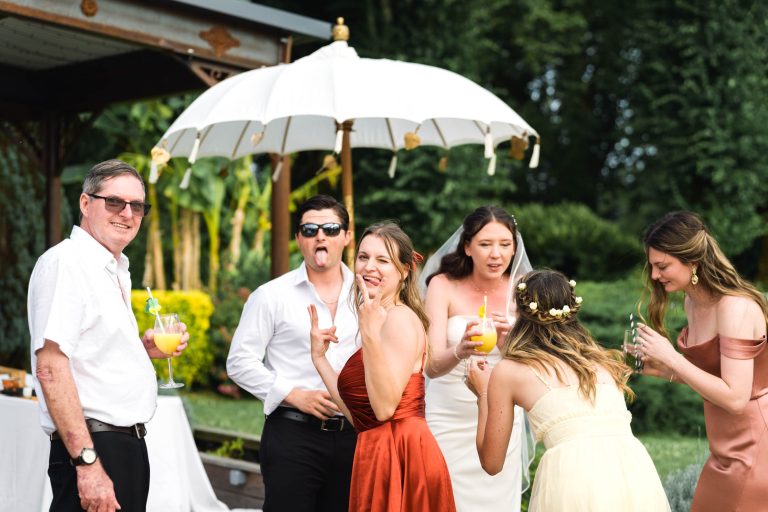 Un groupe de six adultes se tient à l'extérieur, près d'un parasol blanc. Quatre femmes en robe et deux hommes en chemise sourient et rient en tenant des boissons. Un homme fait une grimace ; de grands arbres et un belvédère se trouvent à l'arrière-plan. L'ambiance est joyeuse.