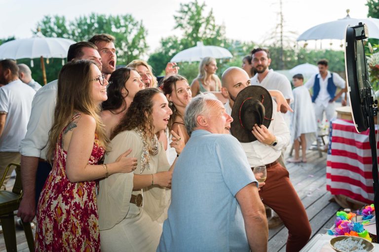 Un groupe de personnes sourit et pose ensemble pour une photo lors d'un événement en plein air. Certains tiennent des boissons et un homme s'amuse à tenir un chapeau. Ils se tiennent près d'un photomaton. Des vêtements d'été brillants et des parapluies blancs sont visibles à l'arrière-plan.