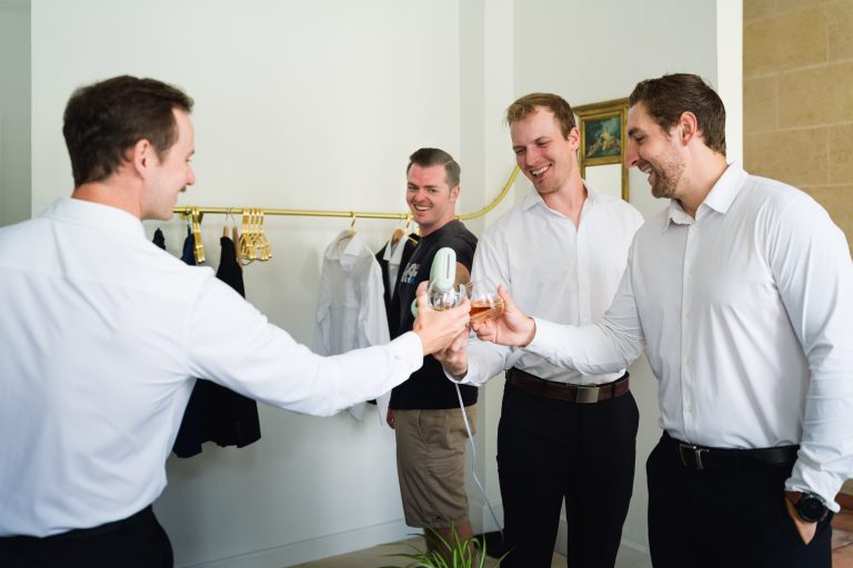 Quatre hommes, trois en chemise blanche et pantalon sombre, et un en tenue décontractée, se tiennent à l'intérieur en souriant. Trois d'entre eux lèvent leur verre pour porter un toast, tandis que l'homme en tenue décontractée observe la scène. Un porte-vêtements doré avec des cintres et des vestes se trouve à l'arrière-plan.