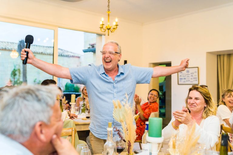 Un homme âgé et joyeux se tient debout, les bras tendus, tenant un micro et souriant largement tandis que les personnes assises autour de lui applaudissent et rient dans une salle lumineuse et décorée de grandes fenêtres et d'un lustre.