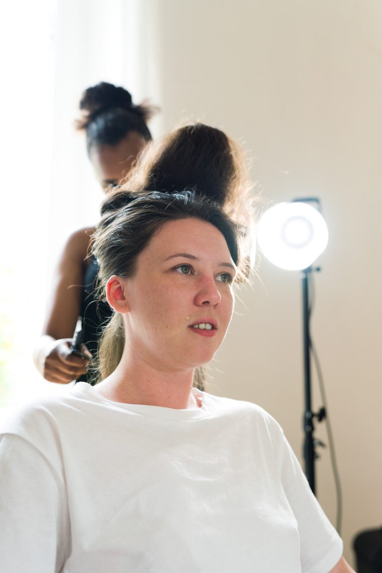 Une femme à la peau claire et aux cheveux bruns est assise sur une chaise, vêtue d'une chemise blanche, tandis qu'une autre femme la coiffe. Un anneau lumineux se trouve à l'arrière-plan, éclairant la scène. La styliste est légèrement floue derrière la femme assise.