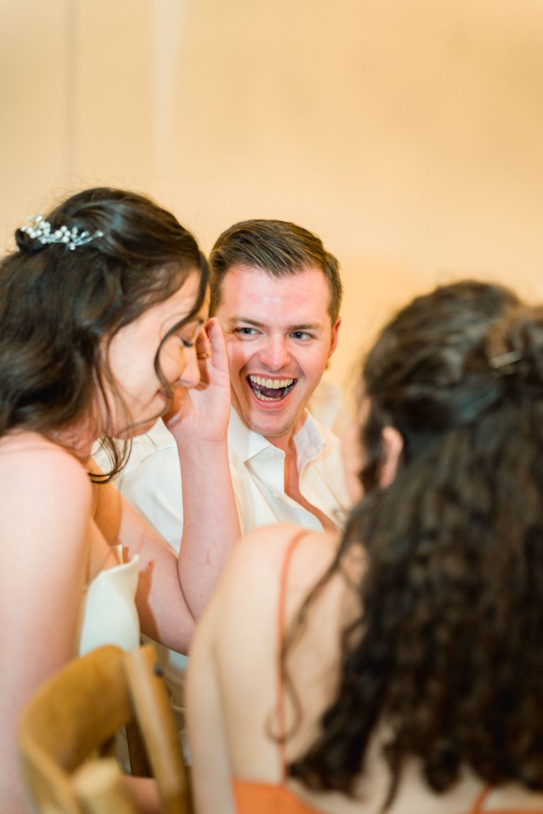 Un homme en chemise blanche rit joyeusement, face à deux femmes aux longs cheveux bruns. L'une d'elles, qui porte une barrette, se couvre le visage de la main tout en souriant. L'arrière-plan est légèrement flou, ce qui crée une atmosphère chaleureuse et joyeuse.