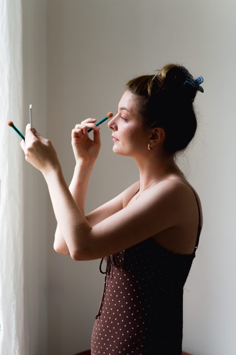 Une femme se tient de profil près d'une fenêtre, vêtue d'une robe brune à pois. Elle tient un petit miroir dans une main et se maquille le visage avec un pinceau dans l'autre. Ses cheveux sont attachés avec une pince et une lumière douce l'éclaire sur le côté.