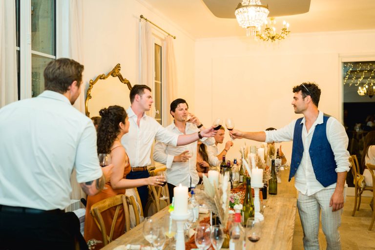 Un groupe de personnes, vêtues de tenues d'été semi-formelles, se lèvent et s'assoient autour d'une longue table à manger, levant leurs verres pour porter un toast. La table est décorée de bougies et de fleurs. Un éclairage intérieur chaleureux et un lustre créent une atmosphère festive et élégante.