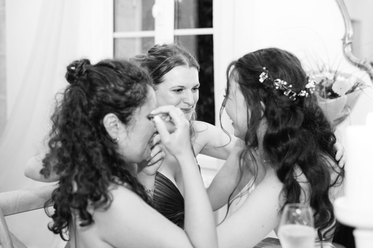 Trois femmes en robe de soirée sont assises l'une près de l'autre, souriant et partageant un moment d'émotion. L'une d'entre elles s'essuie les yeux, peut-être en pleurant de joie. La scène est candide, se déroule à l'intérieur avec un éclairage doux et un arrière-plan flou, suggérant une célébration ou un événement spécial.