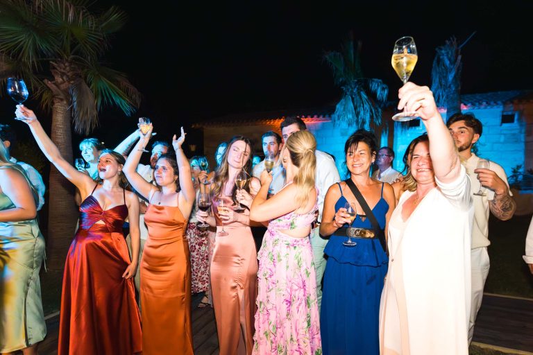 Un groupe de personnes vêtues de tenues colorées et élégantes se tient à l'extérieur, la nuit, levant leurs verres et souriant en signe de célébration. Des palmiers et un éclairage festif créent une atmosphère de fête. Certaines personnes sont en train de se réjouir, tandis que d'autres posent joyeusement devant l'appareil photo.