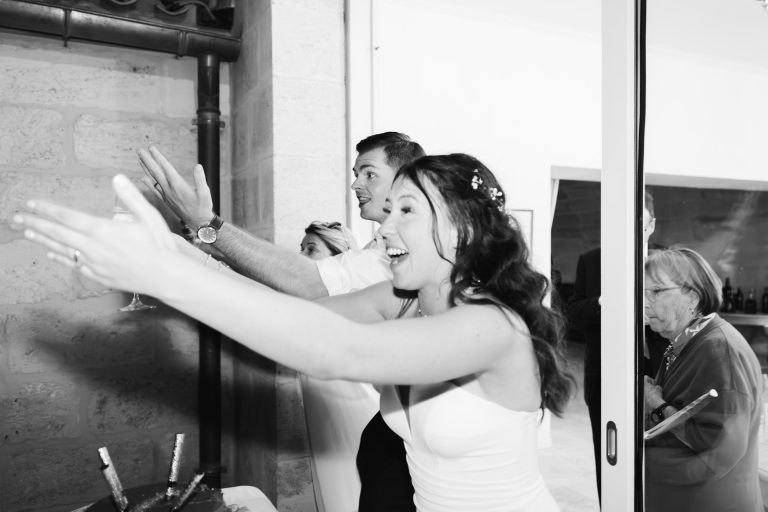 Une femme joyeuse en robe blanche sourit, les bras tendus, à côté d'un homme en chemise blanche qui tend également les bras. Une femme portant des lunettes et un tailleur apparaît à l'arrière-plan, partiellement coupée, à côté d'une fenêtre et d'un mur de pierre.