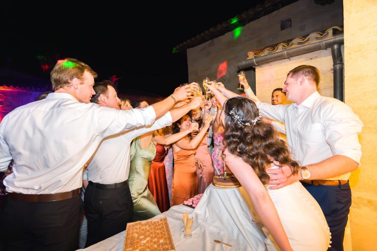 Un groupe de personnes en tenue de soirée, dont une mariée en robe blanche, se réunit autour d'une table pour trinquer avec des flûtes à champagne lors d'une fête nocturne en plein air. La scène est joyeuse, avec des lumières chaudes et un gâteau au premier plan.