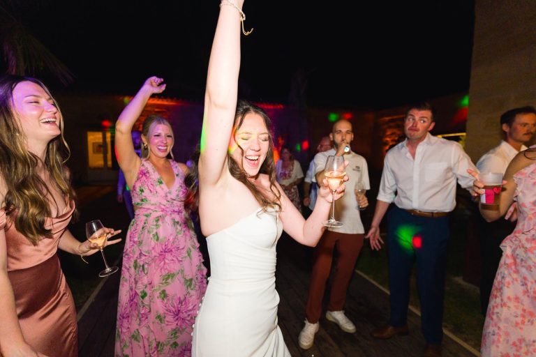 Une femme joyeuse vêtue d'une robe blanche danse, un bras levé, en tenant un verre, entourée de personnes rieuses et souriantes vêtues de tenues colorées, lors d'une fête nocturne animée à l'éclairage vibrant.