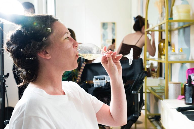 Une femme vêtue d'un chemisier blanc et de bigoudis est assise dans une pièce lumineuse et boit un verre de vin. Elle est photographiée de profil, souriante, près d'un meuble avec des produits de beauté et un miroir. Une autre personne est visible, floue, à l'arrière-plan.