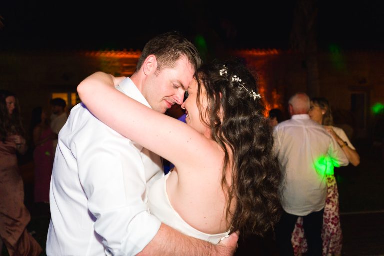 Une mariée et un marié, les yeux fermés et le sourire aux lèvres, s'étreignent en dansant la nuit. La mariée porte une robe bustier et une coiffure florale ; le marié est vêtu d'une chemise blanche. Des lumières colorées et d'autres invités apparaissent doucement à l'arrière-plan.