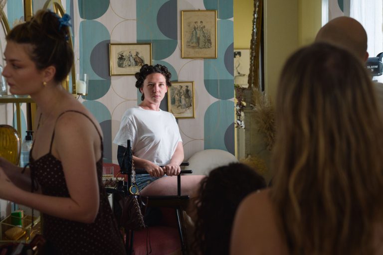 Une femme en chemise blanche avec des bigoudis est assise sur une chaise et regarde devant elle. Elle est entourée de personnes, dont certaines sont floues au premier plan. La pièce est décorée de façon rétro, avec du papier peint à motifs et des œuvres d'art encadrées au mur. L'atmosphère semble chargée.