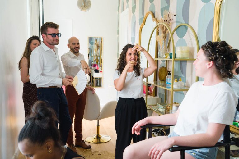 Une femme aux cheveux bouclés est assise, souriante, dans un fauteuil de salon, vêtue d'un tee-shirt blanc. Quatre personnes se tiennent autour d'elle, discutant et riant. Une femme vaporise ses cheveux, tandis que deux hommes en chemise blanche et une femme en robe marron observent, créant une atmosphère animée.
