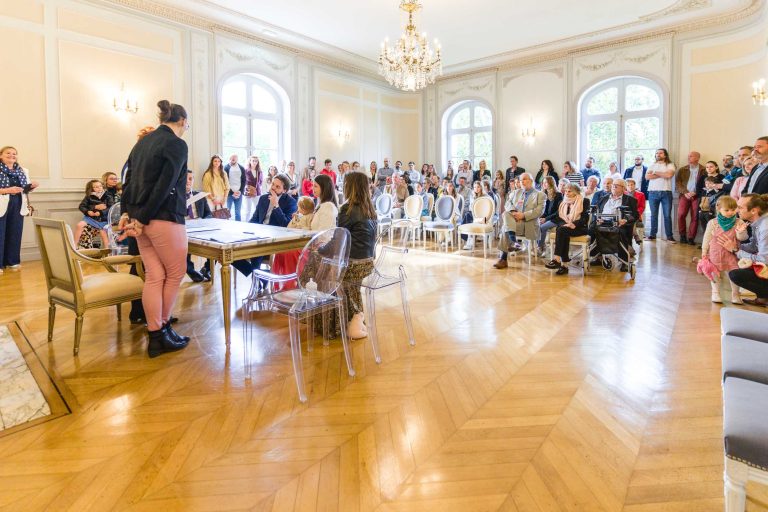 Un grand groupe de personnes, adultes et enfants, est rassemblé dans une pièce lumineuse et élégante, avec de hauts plafonds, des lustres, de grandes fenêtres et du parquet. Certaines personnes sont assises à une table centrale, tandis que d'autres sont debout, ce qui suggère un événement ou une cérémonie formelle.