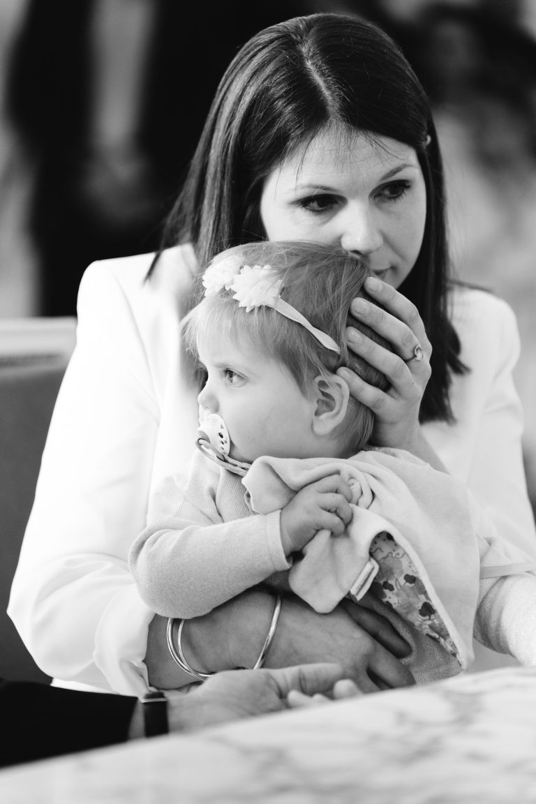 Une femme vêtue d'une veste blanche embrasse affectueusement un jeune enfant avec une tétine et un bandeau fleuri. La femme tient doucement l'enfant contre elle, sa main reposant de manière protectrice sur la tête de l'enfant. La scène est tendre, capturée en noir et blanc.