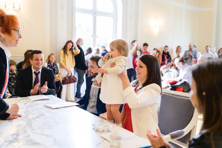 Une femme souriante tient un enfant en bas âge debout sur une chaise dans une pièce lumineuse et élégante remplie de monde. L'enfant porte un vêtement blanc. D'autres adultes s'assoient et regardent, certains souriant. Une femme aux cheveux roux se tient devant une table en marbre, officiant peut-être une cérémonie.