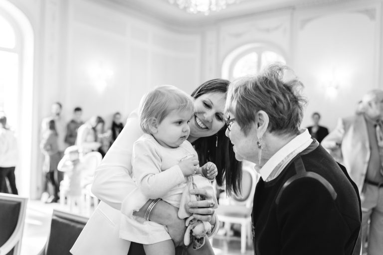Une femme tient un enfant en bas âge tout en souriant à une femme plus âgée, qui se penche en avant pour discuter avec l'enfant. Elles se trouvent dans une pièce lumineuse et élégante avec de grandes fenêtres. Plusieurs personnes sont floues à l'arrière-plan, ce qui donne une atmosphère vivante et sociale.