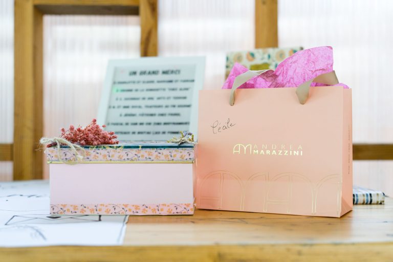 Une boîte décorative contenant des fleurs roses séchées à côté d'un sac cadeau rose Andrea Marazzini rempli de papier de soie rose se trouve sur une table en bois. Un panneau de remerciement et un cadre en bois se trouvent sur un arrière-plan flou.