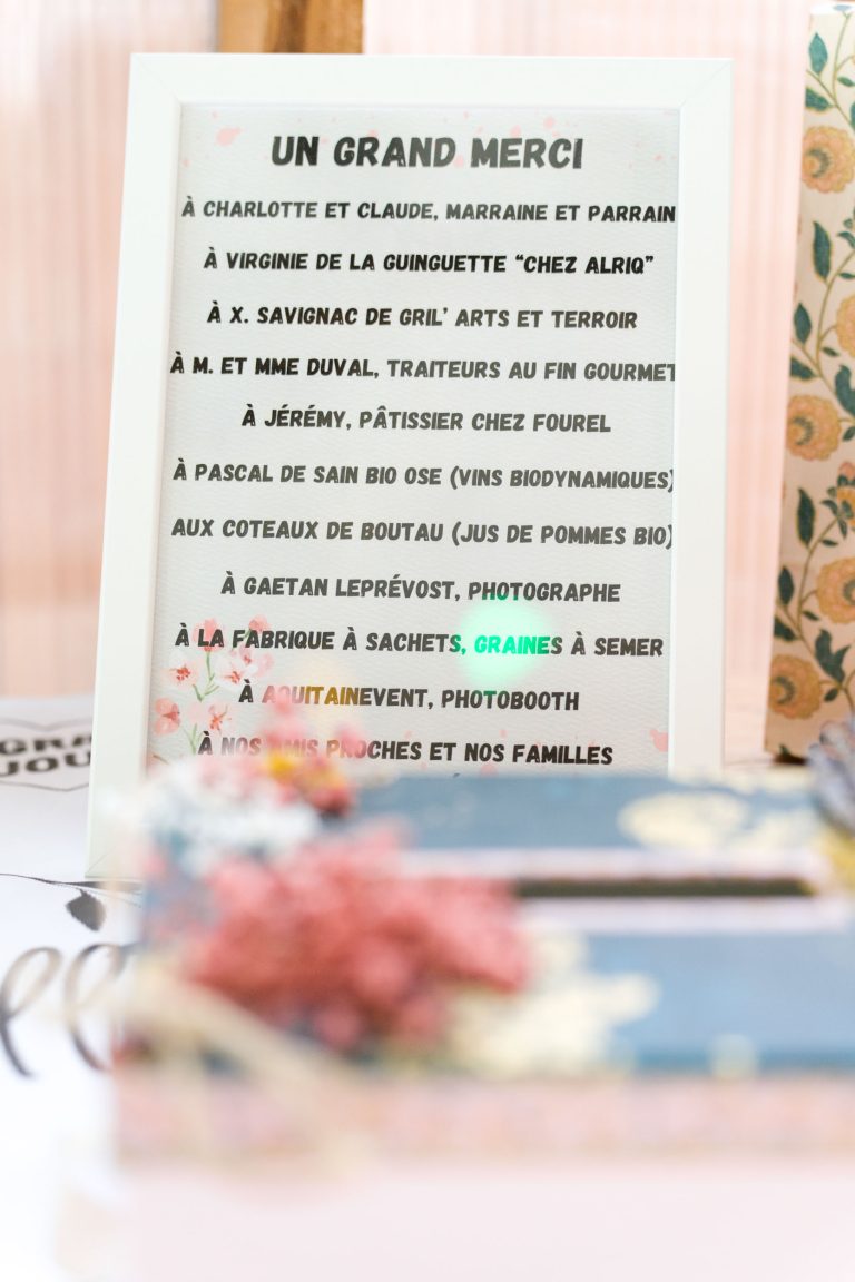Un panneau de remerciement français encadré énumère les noms et les professions de personnes et d'entreprises, telles que les traiteurs, les boulangers, les photographes, etc. Le cadre est posé sur une table avec, au premier plan, une boîte à fleurs bleue floue et des décorations roses.