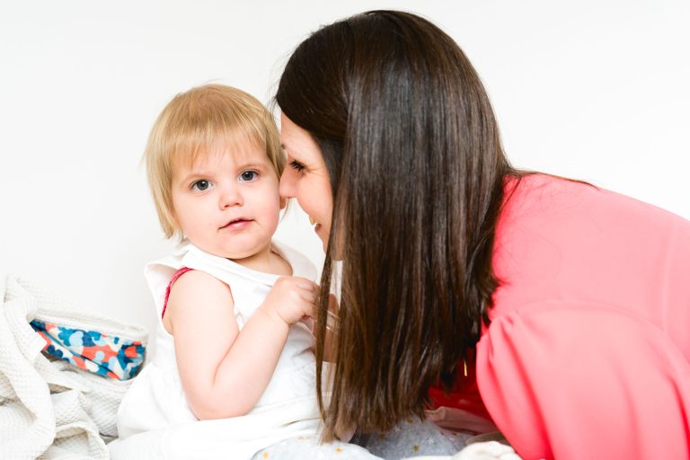 Un bambin aux cheveux blonds vêtu d'un ensemble blanc est assis sur un lit et regarde l'appareil photo, tandis qu'une femme aux longs cheveux bruns vêtue d'une chemise rose se penche vers lui et lui sourit, créant ainsi un moment de chaleur et d'affection. Une couverture blanche à motifs bleus et rouges se trouve à proximité.